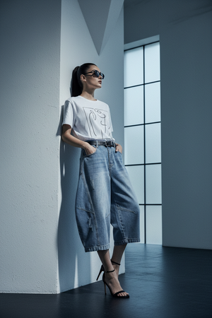A woman stands in a minimalist room with white walls and a large window, wearing a white graphic t-shirt, wide-leg light-wash denim culottes, and black high-heeled sandals.